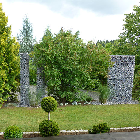 Ein Gartengrundstück mit modernen Steinwänden und Steinkörben aus Gabionen, drumherum verschiedene Pflanzen und Bäume